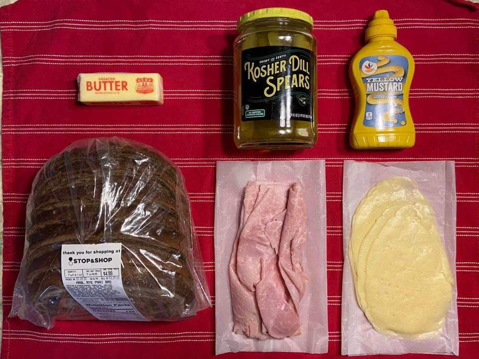 Flat lay of dark rye bread, sliced ham, sliced Swiss, a jar of pickles, mustard, and a stick of butter.