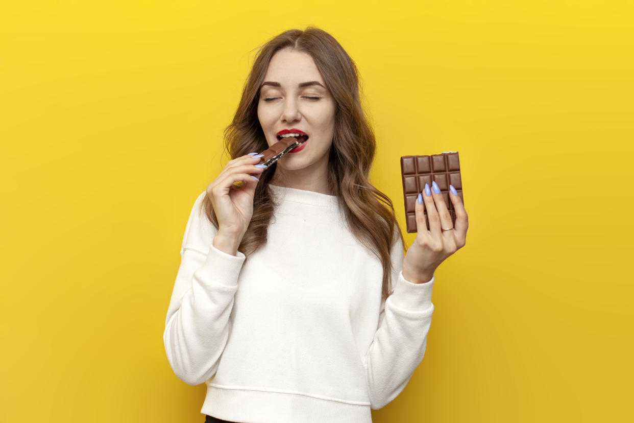 young girl eats and bites big chocolate bar foods and smiles and rejoices on yellow isolated background, happy woman with sweet meal shows toothy smile