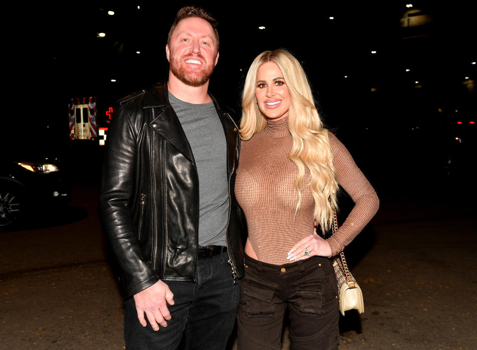 Kroy Biermann wears a grey tshirt and black jacket next to Kim Zolciak who's wearing a tan shirt with black pants.