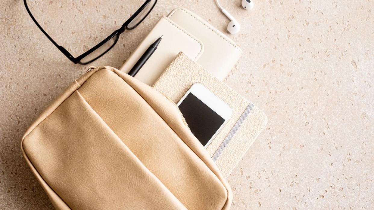 beige woman handbag with purse, note pad, pen, smart phone, eyeglasses and in ear headphones on concrete background from above, top view