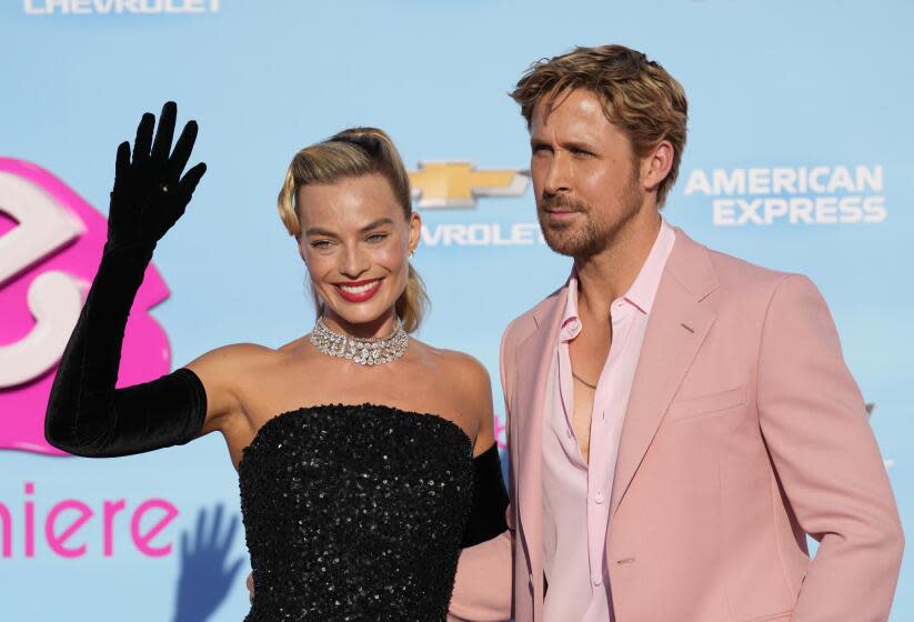 Margot Robbie is wearing a black dress and is smiling and waving next to Ryan Gosling who is in a pink suit