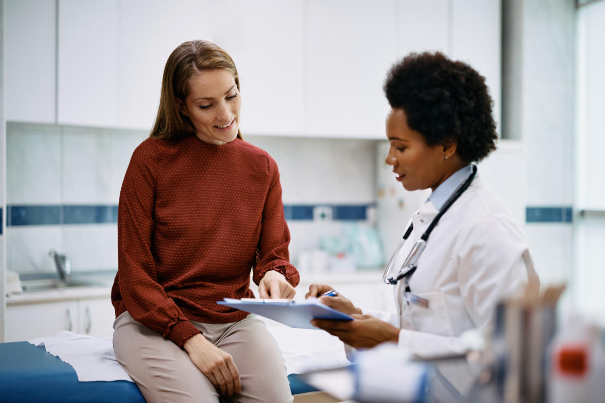 A female patient discusses formalities with a female doctor holding a clipboard with holding documents.