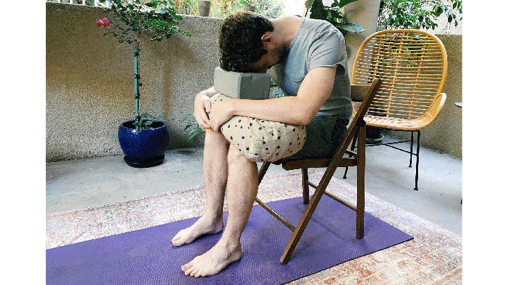 Man seated on a chair draping his body forward over a bolster with his forehead resting on a block in the yoga pose known as Child's Pose