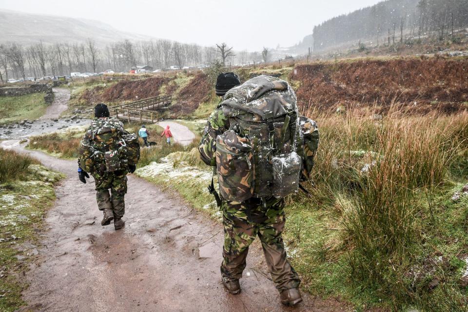 Two soldiers wearing camouflage and carrying large packs walk down a muddy and snowy mountain path.