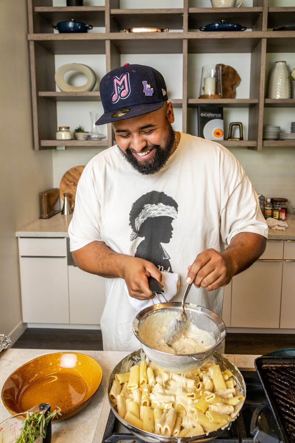 A man in a ball cap and wearing a T-shirt that says "Black Pot" smiles as he poses for a photo behind a gas range.