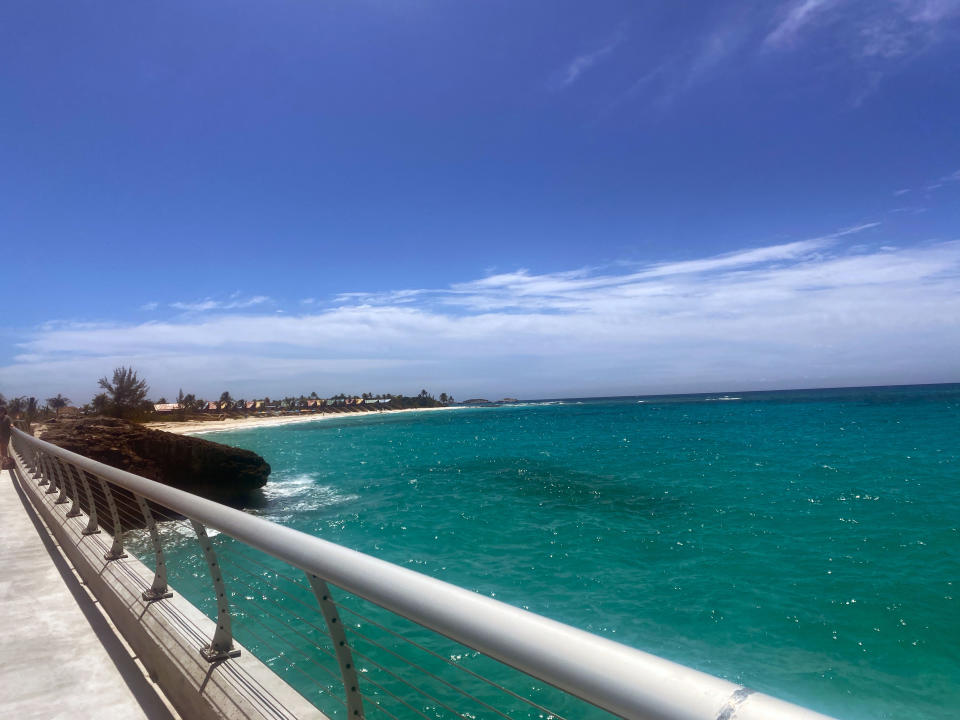 A walkway with railings overlooks a calm, teal ocean adjacent to a sandy beach with distant houses and trees under a clear sky