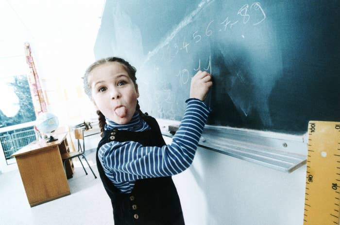 A young girl at a classroom chalkboard playfully sticks her tongue out while writing numbers. Chairs and a desk are visible in the background