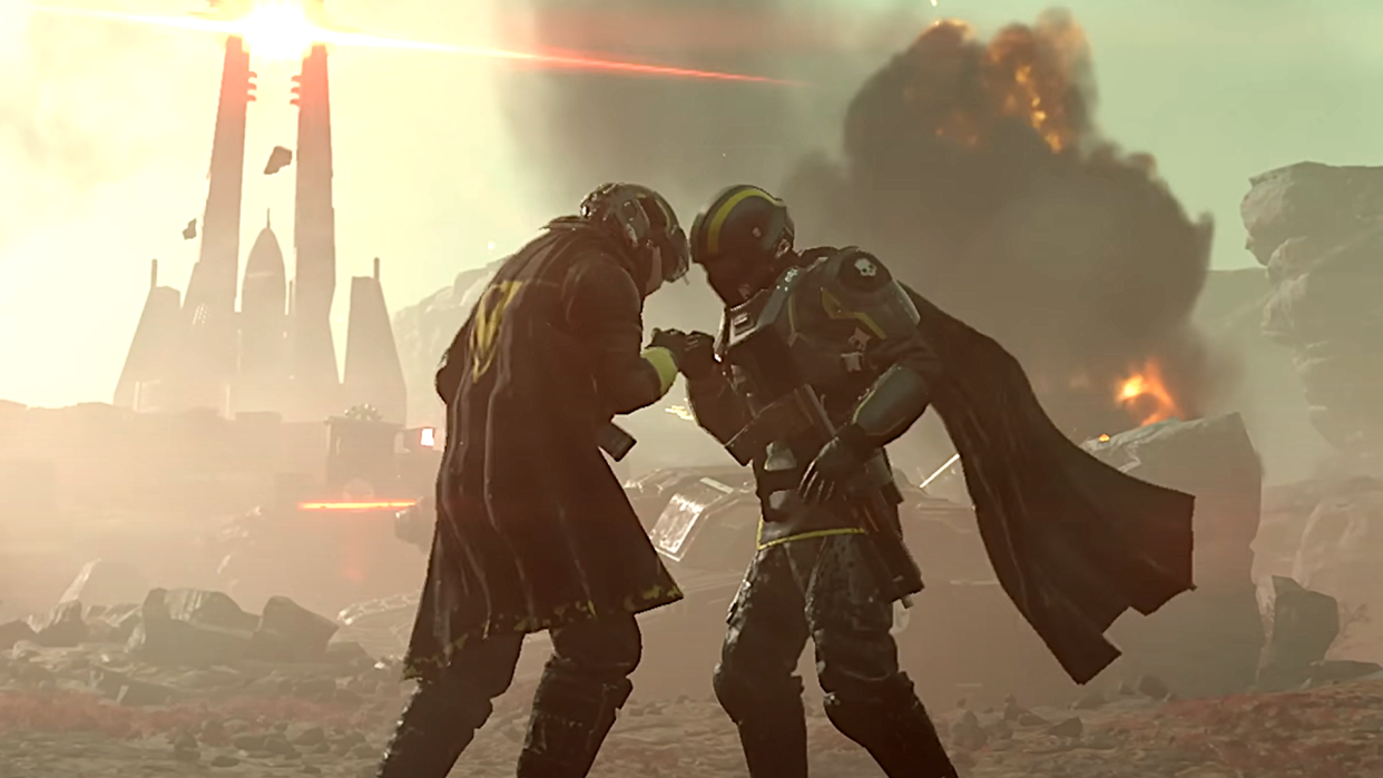  Two Helldivers share a tender moment in Helldivers 2, while Automatons ravage the landscape behind them with explosions. 