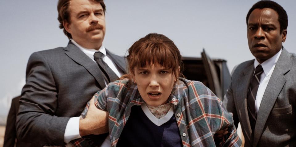 Two men in suits hold the arms of a teenage girl with brown hair, bangs, and wearing a plaid shirt (the character Eleven, played by Millie Bobby Brown).