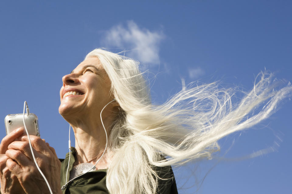 Older woman with long white hair smiling, listening to music on an iPod with earphones, wind blowing her hair against a clear blue sky