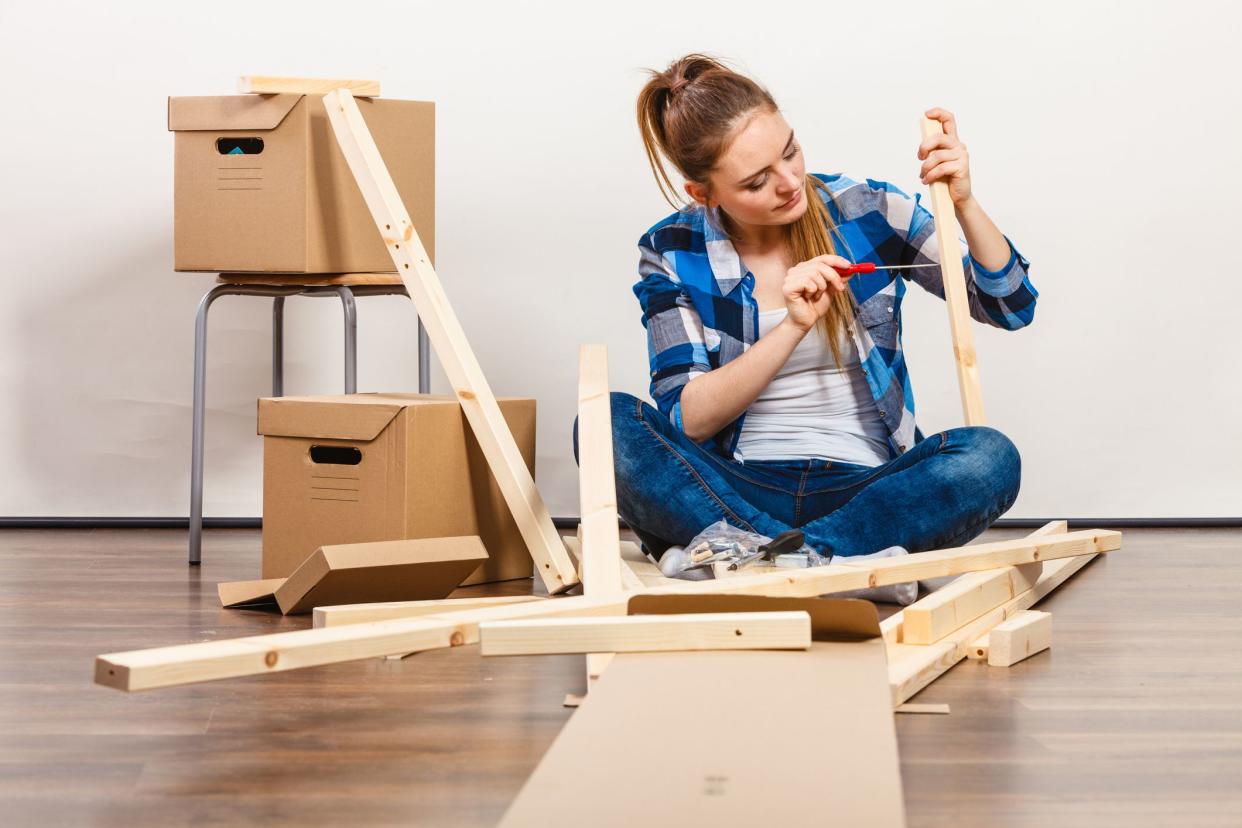 Young woman sitting on the floor putting together furniture, surrounded by wooden furniture parts and two boxes, a white wall in the background