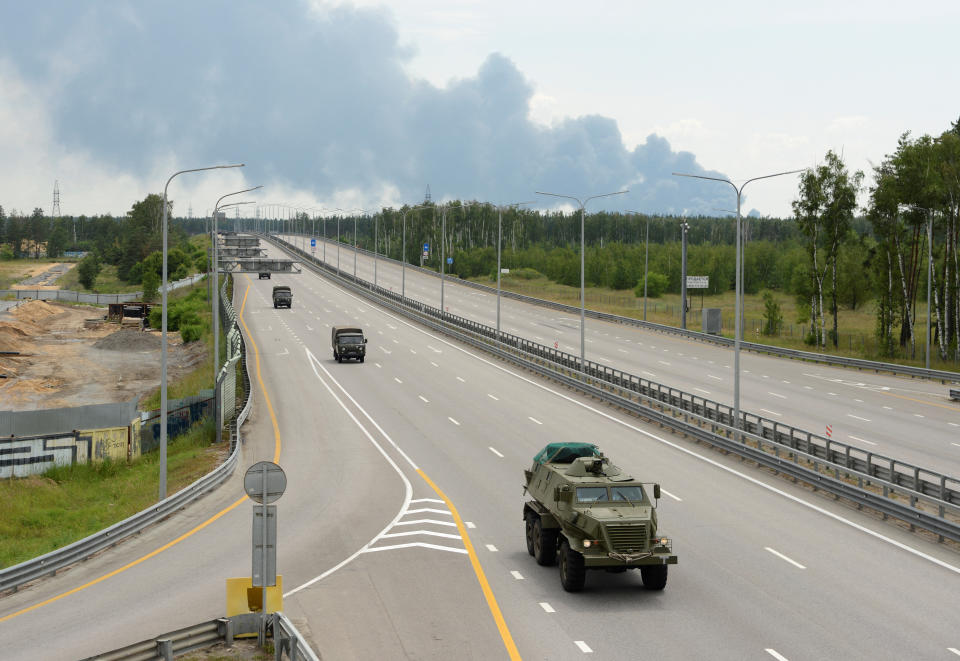 A half dozen military vehicles, in a column spaced with about 100 yards between them, drive along what appears to be a closed highway, with trees and smoke in the background.