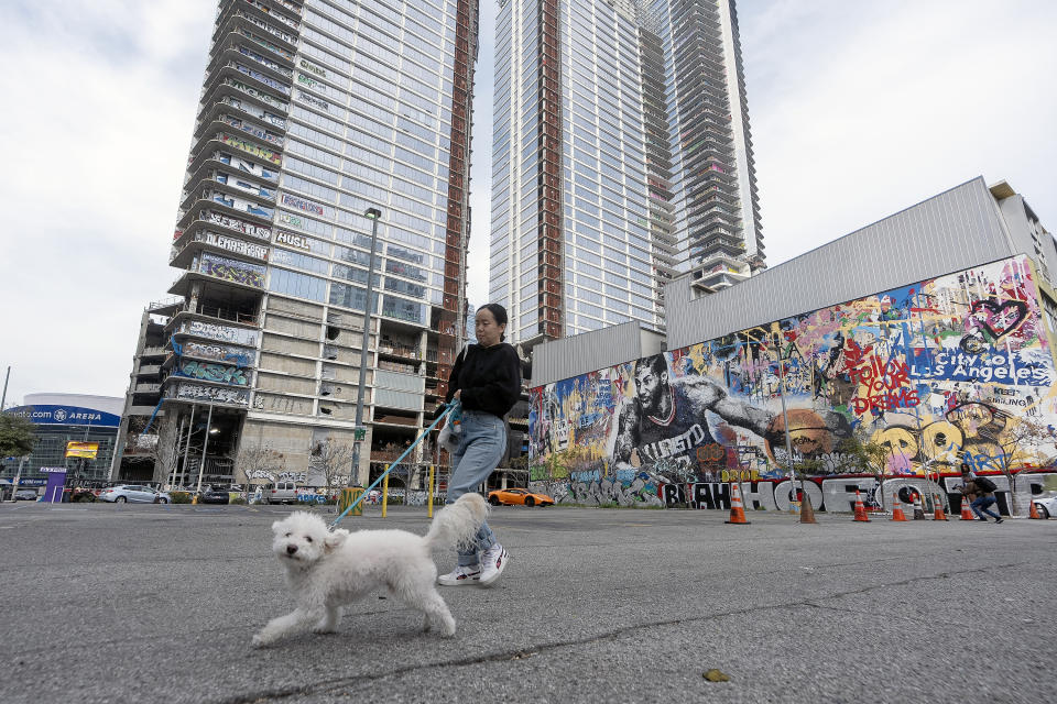 A local resident walks her dog, past an unfinished complex of high-rise towers that have recently been vandalized with graffiti and used for dangerous social media stunts after the developer ran out of money is seen in downtown Los Angeles Friday, Feb. 16, 2024. The three towers have become an embarrassment in a high-profile area that includes Crypto.com Arena, home of major sports teams and events such as the Grammys, as well as the Los Angeles Convention Center and the L.A. Live dining and events complex. (AP Photo/Damian Dovarganes)