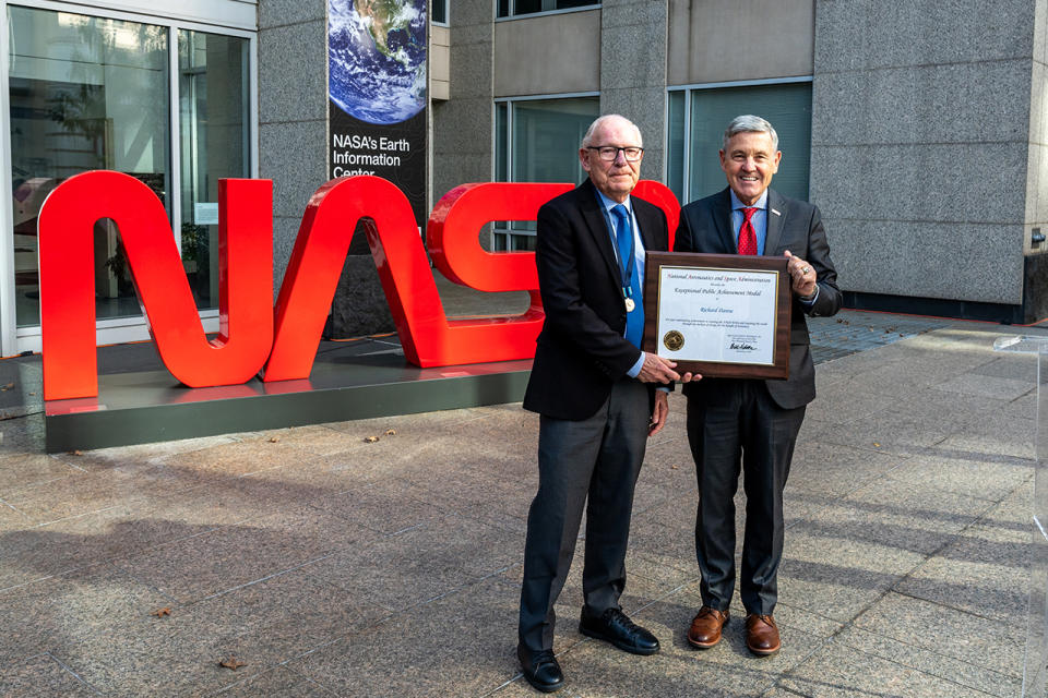 two smiling older men in suits hold a framed certificate in an outdoor courtyard.