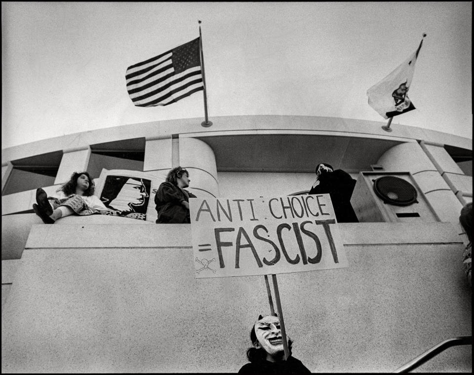 A demonstrator in a demon mask during a rally stands beneath California and US flags with a sign reading "anti-choice equals fascist"