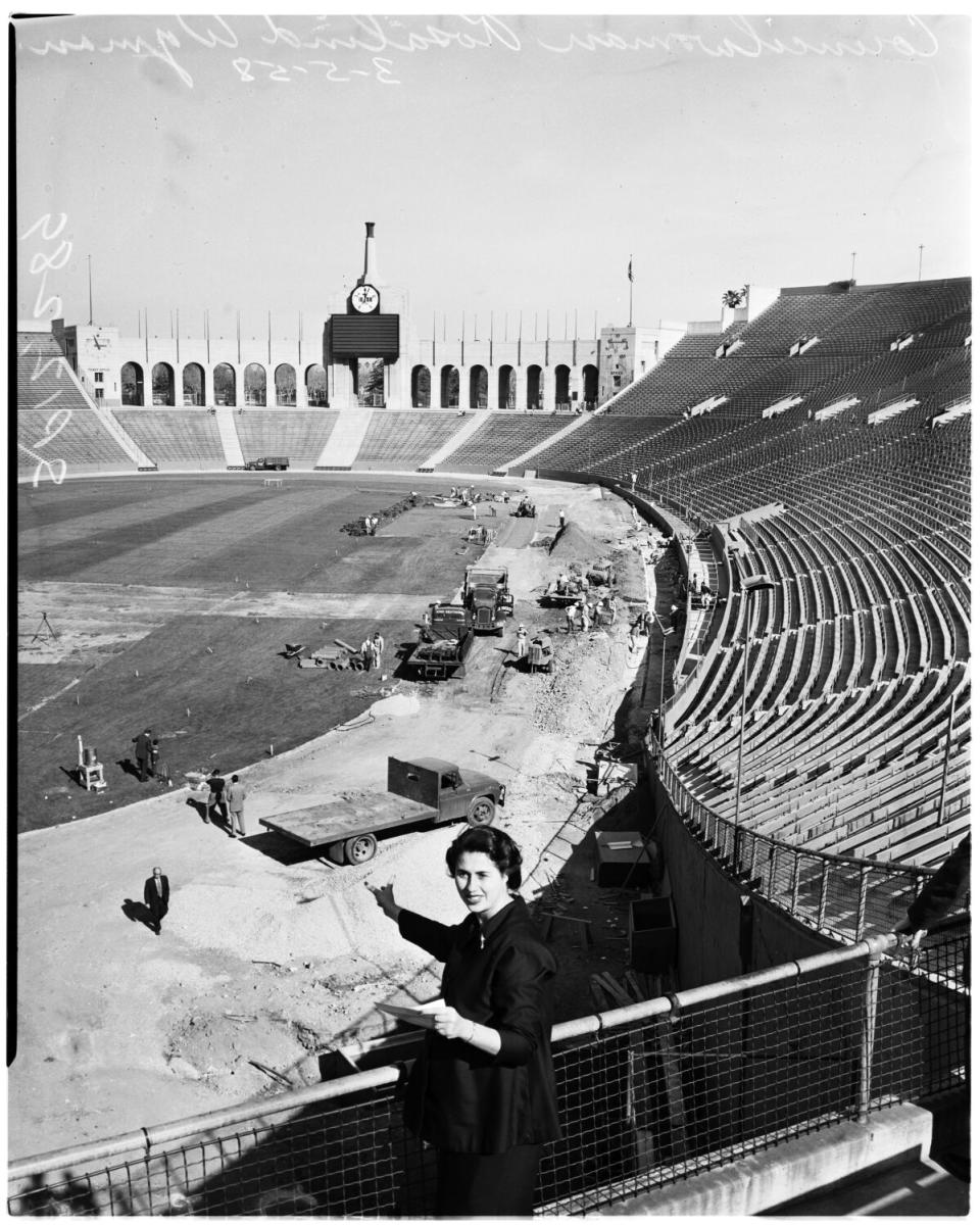 Rosalind Wyman in the stands of a coliseum while workers and vehicles were below