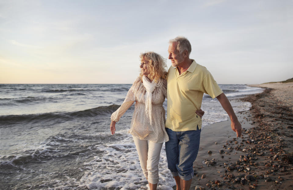 An older couple walks along a sandy beach, smiling and enjoying the ocean view as they hold each other close and stroll by the water