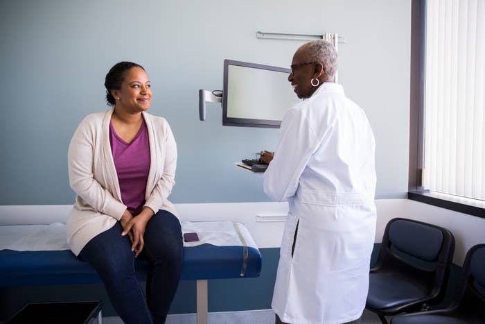 A patient sits on the exam table while conversing with a doctor who holds a clipboard