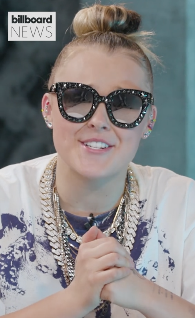 JoJo Siwa wearing oversized, embellished sunglasses and layered necklaces, hands clasped together, smiling