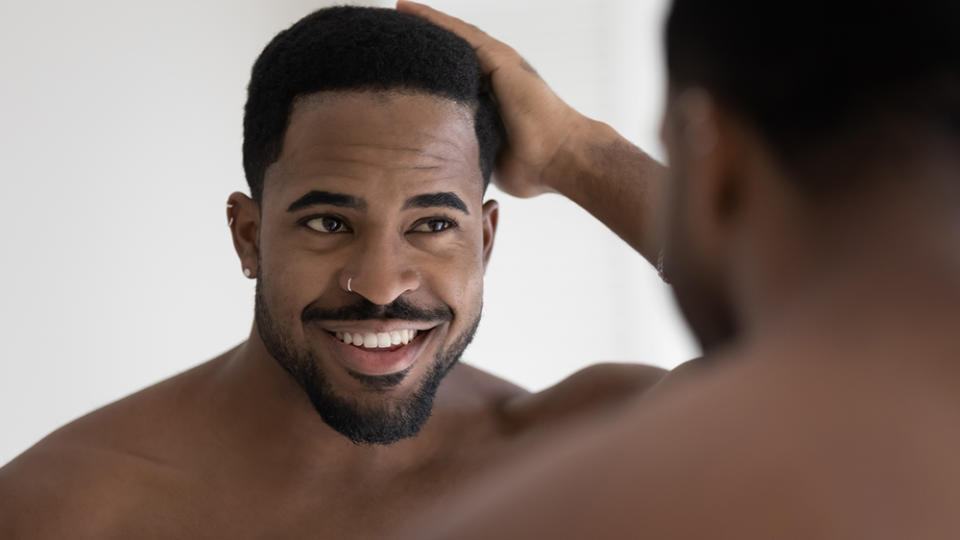 Handsome shirtless muscular build African man standing in bathroom looking in mirror touch his natural afro hair, concept of happy client of barber shop services, haircare treatment, cosmetics for men