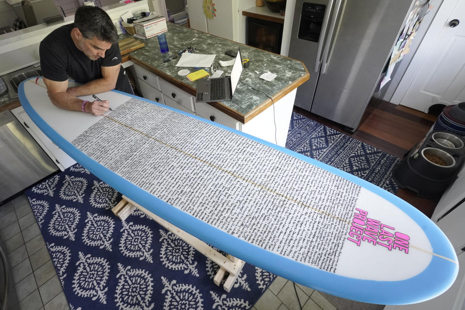 Dan Fischer, of Newport, R.I., uses a paint marker to place names of lost loved ones on one of his surfboards at his home, in Newport, Wednesday, May 25, 2022. Fischer, 42, created the One Last Wave Project in January 2022 to use the healing power of the ocean to help families coping with a loss, as it helped him following the death of his father. Fischer places the names onto his surfboards, then takes the surfboards out into the ocean as a way to memorialize the loved ones in a place that was meaningful to them. (AP Photo/Steven Senne)