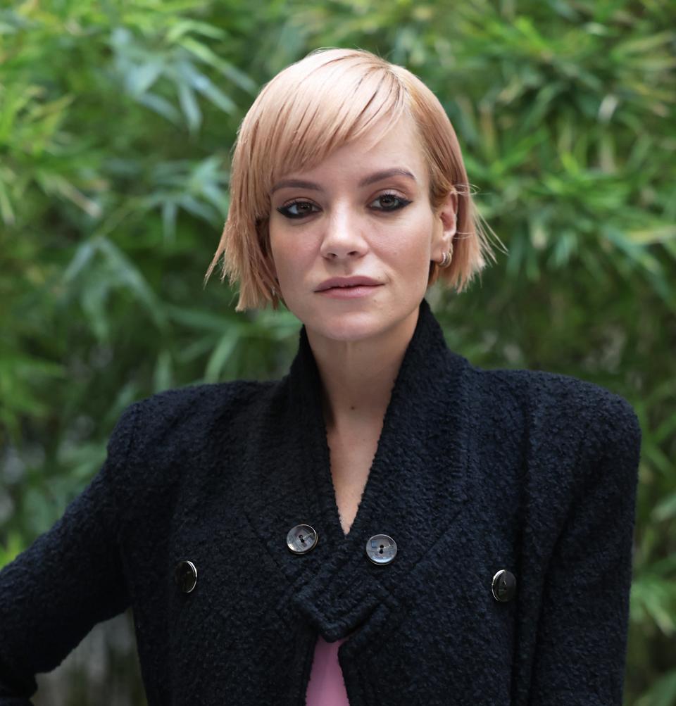 Lily Allen is dressed in a stylish long dark coat over a pink outfit, holding a textured clutch purse. She stands against a backdrop of green foliage