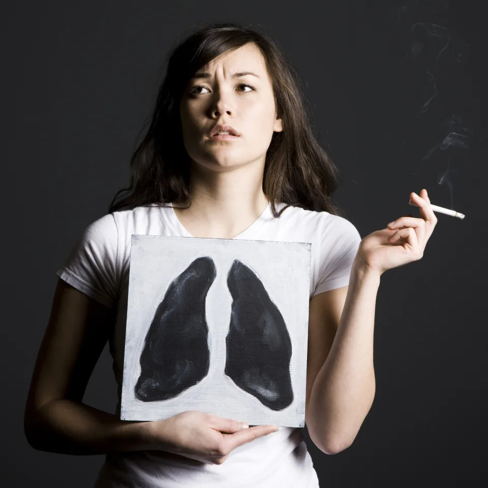 Woman smoking against a black background holding a poster with black lungs on it.