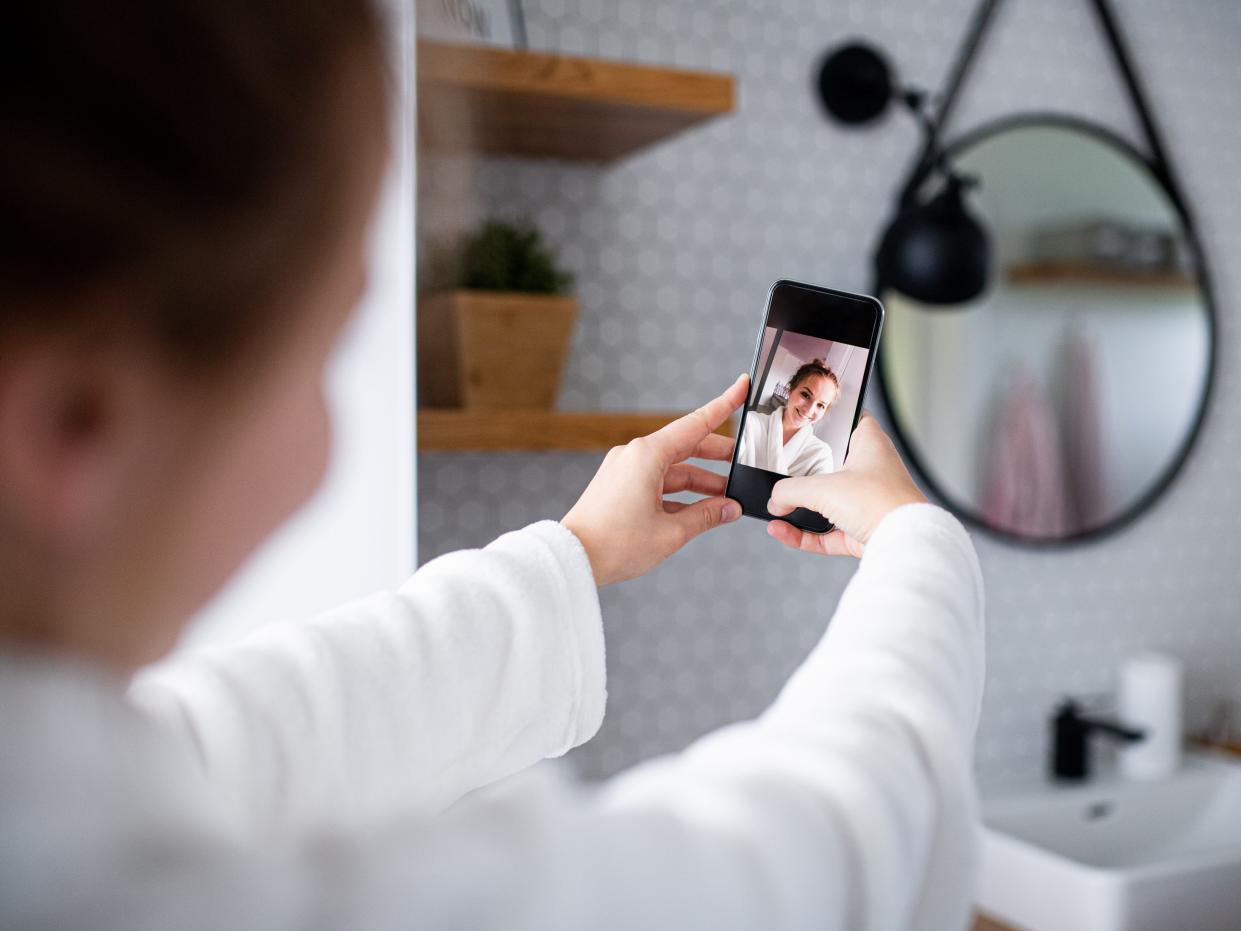 Young woman with bathrobe and smartphone in bathroom, taking selfie