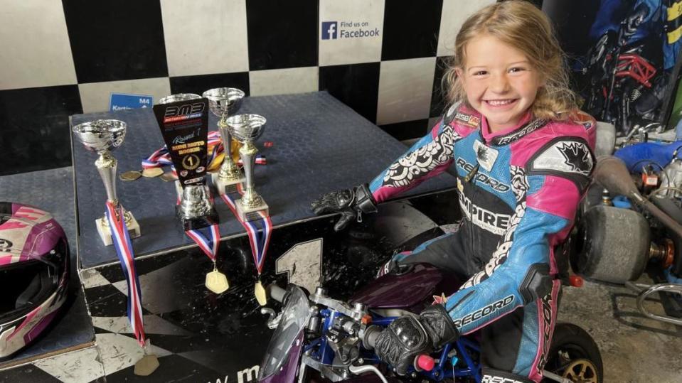 A young blonde girl sitting on a mini-moto bike in a brightly coloured racing suit next to her medals and trophies 