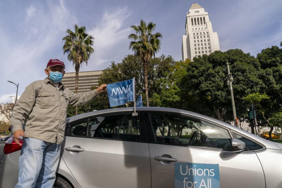 A Uber driver wearing a mask stands beside his car that has a Unions For All banner on the passenger door.