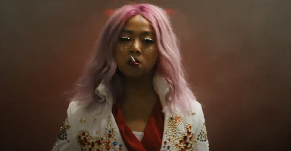 Stephanie Hsu in Everything, Everywhere, All At Once, with pink hair, glitter makeup and a white suit, smoking a cigarette