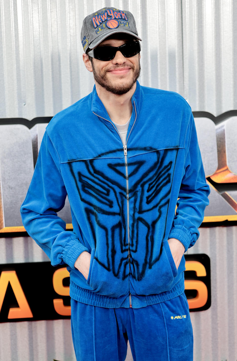 Pete Davidson wearing a blue tracksuit and a cap at a media event, smiling with hands in pockets, standing in front of a metallic background