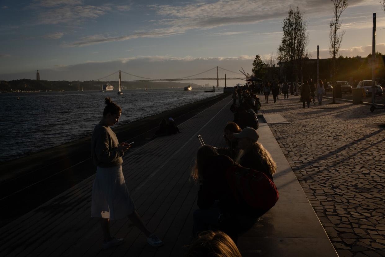 People congregate at dusk by a river, with views of a bridge