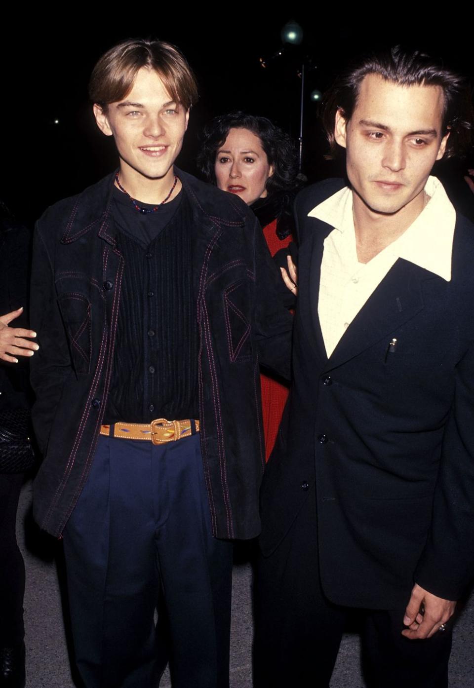 leonardo dicaprio and johnny depp stand next to each other, dicaprio smiles and wears a denim jacket, black vest, black shirt and royal blue pants with a brown belt, depp leans toward the camera and wears a black suit with a white collared shirt