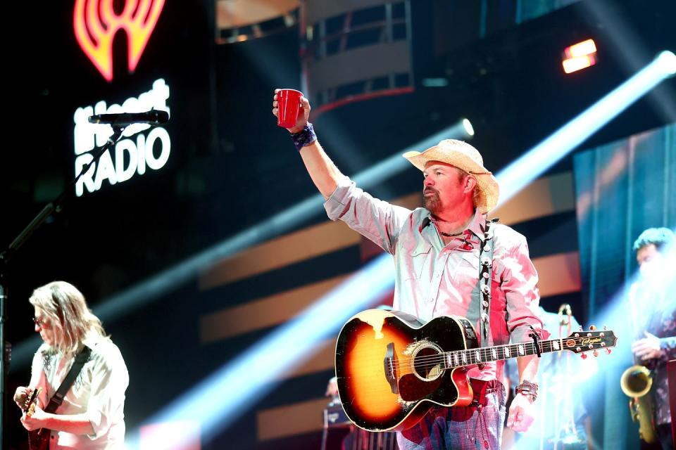 toby keith stands on a stage with a few spotlights shining down, he holds a red solo cup above his head and has an acoustic guitar on a shoulder strap, he wears a straw colored cowboy hat, gray collared shirt and jeans