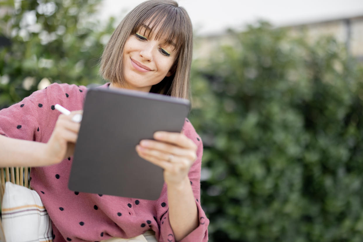 Portrait of young creative woman works with stylus on a digital tablet at backyard outdoors. Concept of remote work or leisure time with gadgets at cozy atmosphere