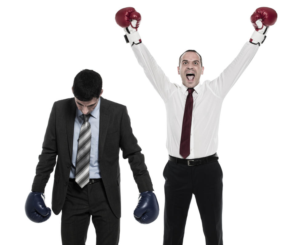 Two men in business attire are wearing boxing gloves. One man hangs his head in defeat, while the other raises his arms in victory.