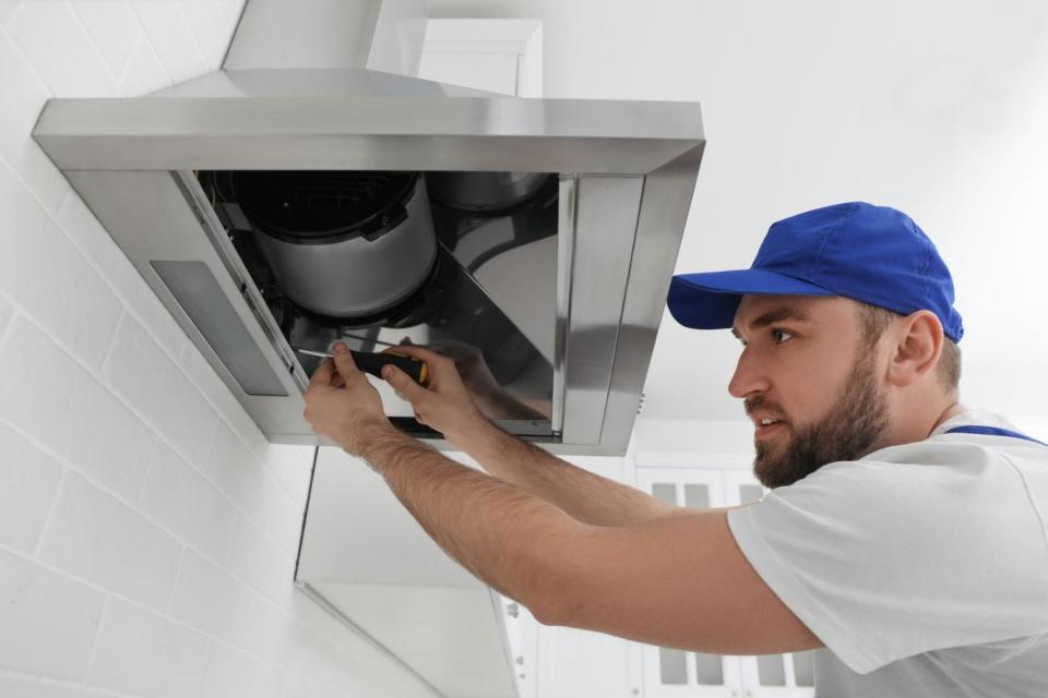 A man in a blue cap screws in a range hood.