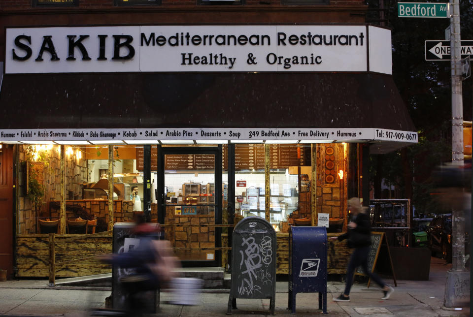 In this Wednesday, Oct. 9, 2019, photo, people rush past Sakib Mediterranean restaurant, co-owned by Syrian refugee Diaa Alhanoun and his partner, in Brooklyn’s Williamsburg neighborhood, in New York. Alhanoun's partner Mohammad Ayasrah is visible through the window, left, speaking on his phone. (AP Photo/Kathy Willens)