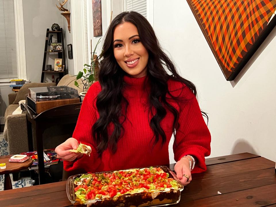 The writer, wearing a red shirt, smiles and holds a tortilla chip with 7-layer dip on it. A casserole dip garnished with tomatoes, olives, and guacamole sits in front of her