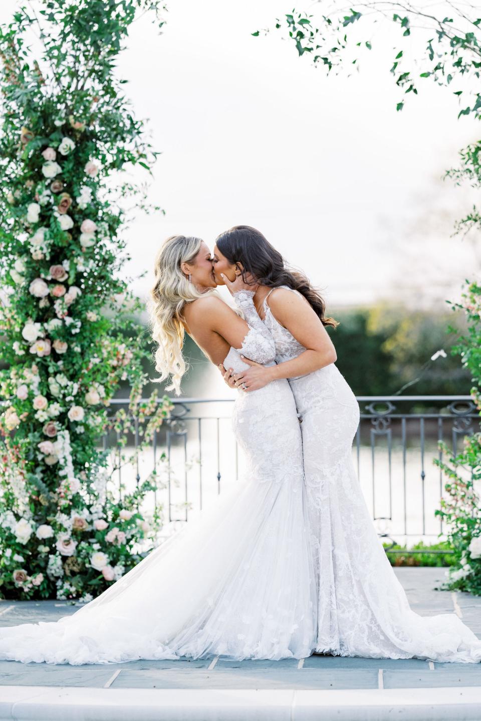 Two brides kiss under a floral archway.