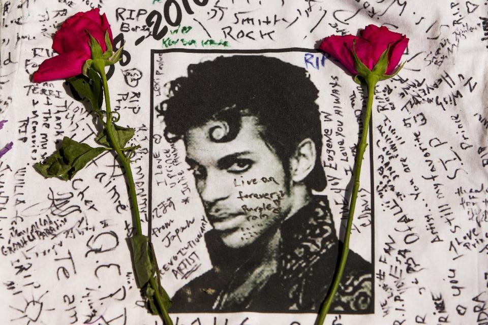 FILE - In this April 22, 2016 file photo, flowers lie on a T-shirt signed by fans at a makeshift memorial for musician Prince outside the Apollo Theater in New York. The singer died April 21, 2016, at the age of 57. With the loss of several icons of Generation X’s youth, the year 2016 has left the generation born between the early 1960s and the early 1980s, wallowing in memories and contemplating its own mortality. (AP Photo/Andres Kudacki, File)
