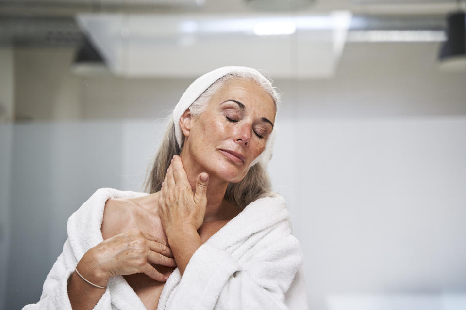 A woman in a bathrobe, with a towel wrapped around her head, touches her neck and appears relaxed with eyes closed