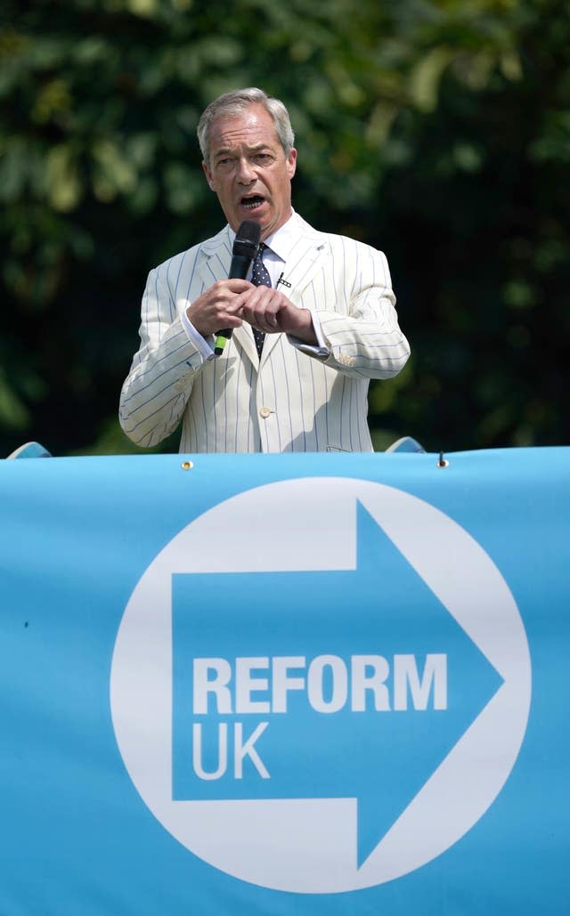 Nigel Farage in pinstriped cream suit jacket speaking into a mic behind a Reform UK banner
