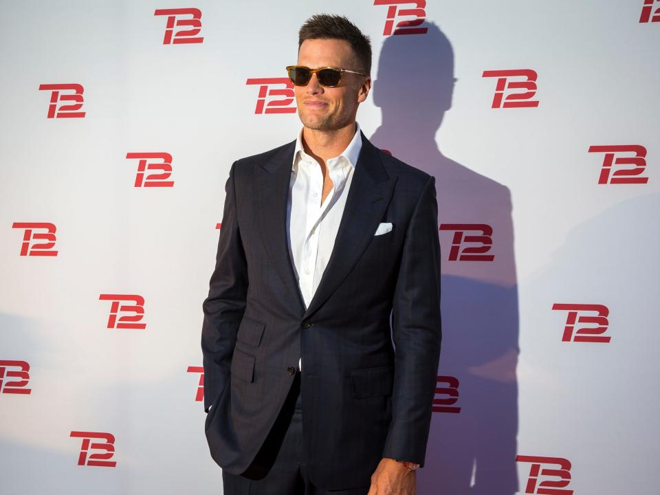 Tom Brady stands in a black suit in front of a backdrop with the TB12 logo.