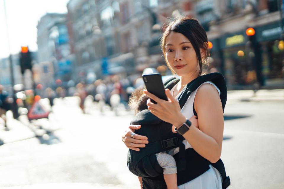 Beautiful Asian mother carrying her baby daughter and ordering taxi service via mobile app on smart phone while walking in the city.