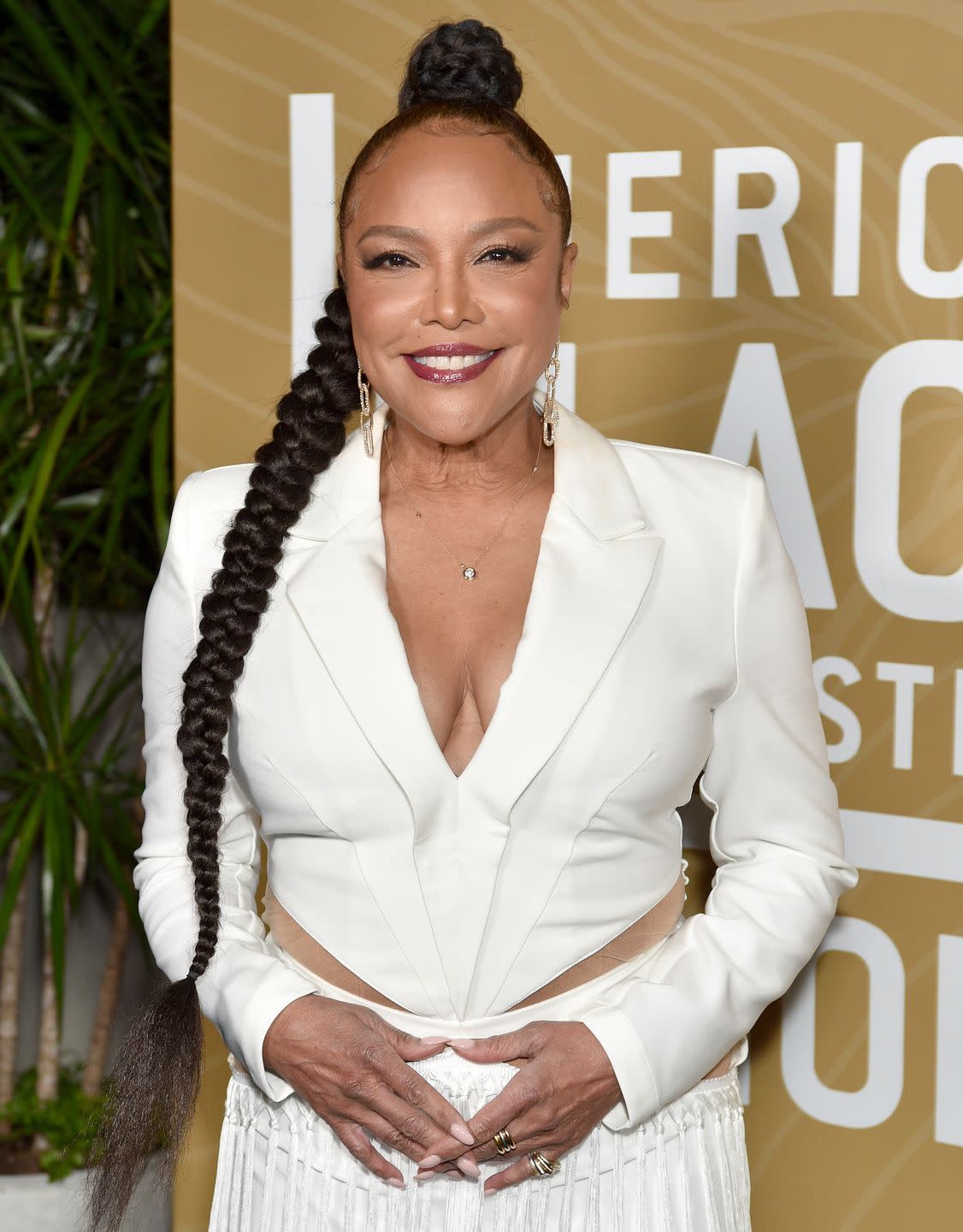 lynn whitfield, a woman stands looking at the camera and smiling, she wears her long black hair in a braid and a white suit top and trousers