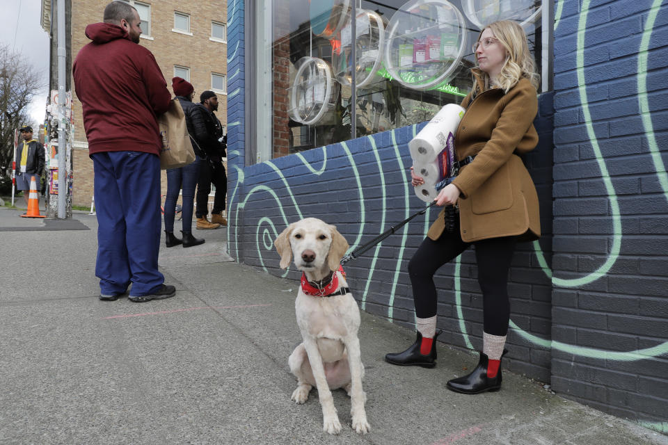 In this photo taken March 24, 2020, Mia Grace, right, holds a package of toilet paper as she and her dog Breezy observe social distancing chalk marks on the sidewalk while waiting to get in to The Reef Capitol Hill, a marijuana store in Seattle, which was limiting the number of people in the store at one time to help slow the spread of the new coronavirus. Earlier in the week, Washington Gov. Jay Inslee ordered nonessential businesses to close and the state's more than 7 million residents to stay home in order to slow the spread of the new coronavirus. In Washington and several other states where marijuana is legal, pot shops and workers in the market's supply chain were deemed essential and allowed to remain open. (AP Photo/Ted S. Warren)