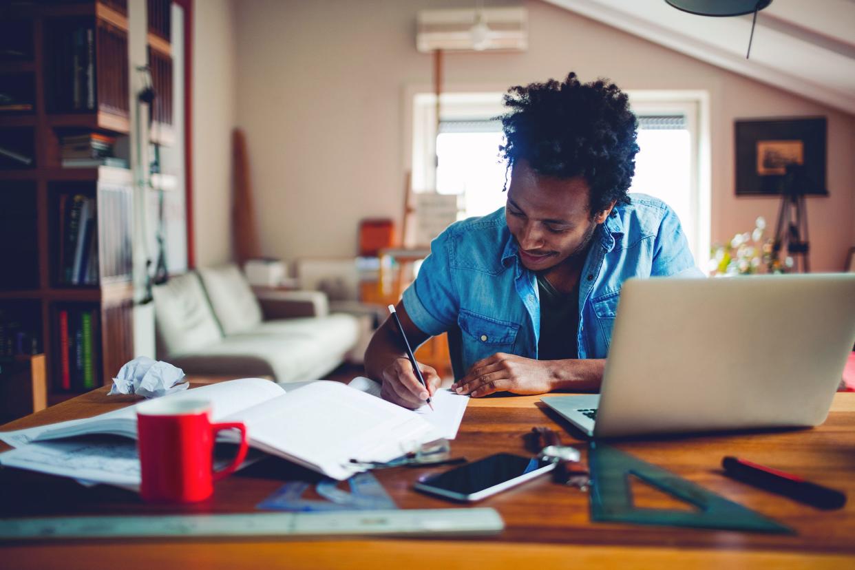 Young male student studying at desk with laptop, textbooks, writing with a pencil on a notebook, mobile, protractor, ruler, and a red coffee cup on desk, with a blurred background of couch and large window in a room at home