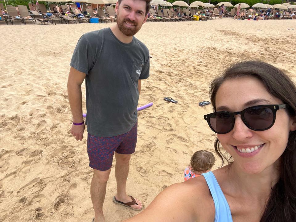 A woman taking a selfie on the beach with her husband and child.